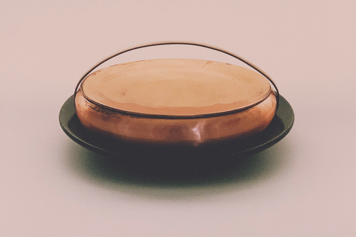 A round copper box with a copper handle on top of a green marble dish on a beige background