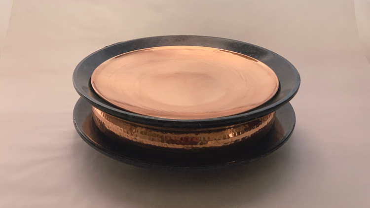 A copper uruli dish with two stone plate made of black granite placed on top and bottom of the dish, placed on the top of the top plate is a copper lid