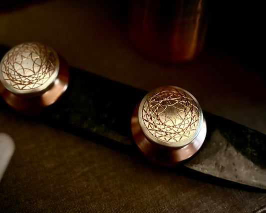Prakasham Vallam are two small round containers made of copper and gilted with silver with etchers patterns  on a dark  green marble stone surface with a textured wall in the background.