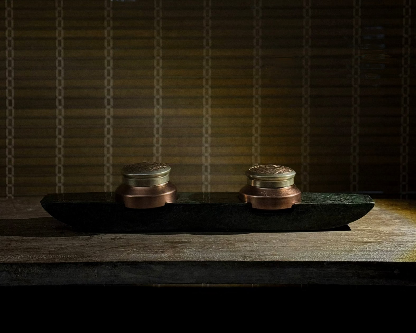 Prakasham Vallam are two small round containers made of copper and gilted with silver with etchers patterns  on a dark  green marble stone surface with a textured wall in the background.