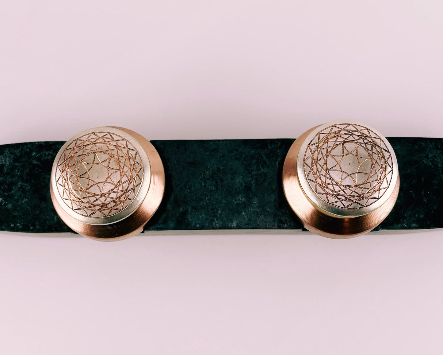 Prakasham Vallam are two small round containers made of copper and gilted with silver with etchers patterns  on a dark  green marble stone surface with a textured wall in the background.